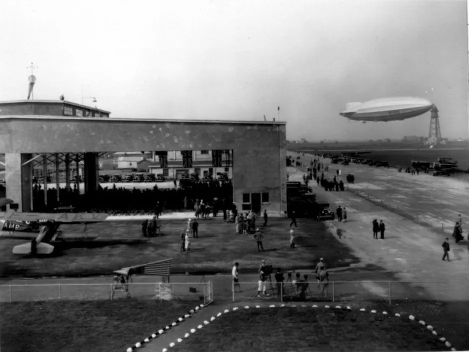 Une photographie noir et blanc montre un hangar d’aéroport et une piste d’atterrissage, ainsi que l’immense dirigeable amarré à une haute structure ressemblant à une tour.