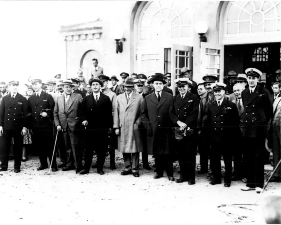 Une photographie noir et blanc montre un groupe d’hommes, certains en uniforme militaire et d’autres en costumes et longs manteaux, prennent la pose à la cérémonie soulignant l’arrivée du dirigeable.