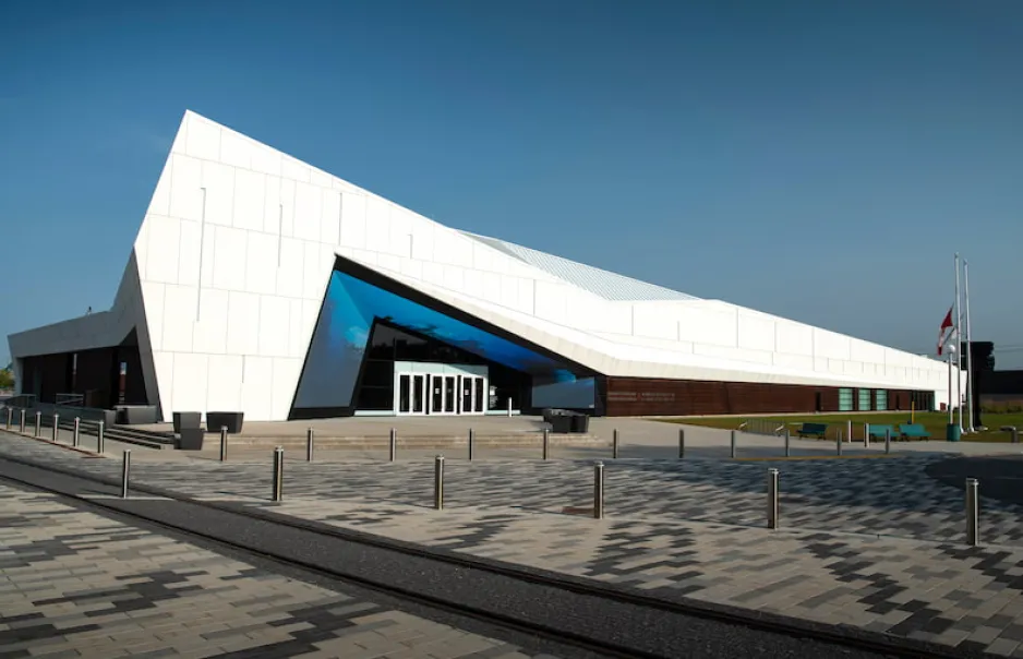 The façade of a wide, geometric white building set against a clear blue sky. Around and above the building’s front doors, a large screen displays vivid blue tones.
