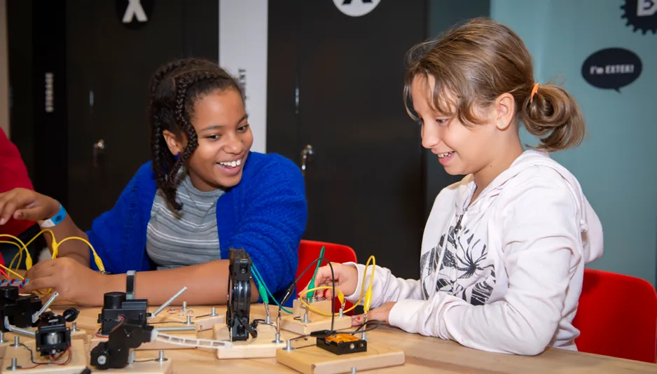 Two girls wide wide smiles sit a table and handle small motors, wires and batteries.  The girl on the left has dark skin and hair and is wearing a striped shirt and blue cardigan.  The girl on the right has an olive complexion and light brown hair and is wearing a white hoodie with a zebra image.