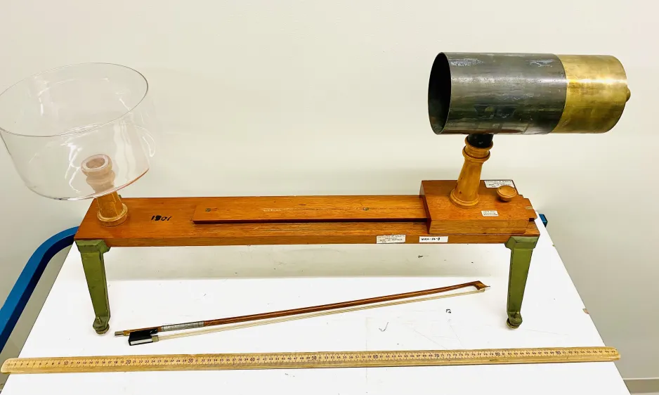 Full frame shot of the cherry bowl resonance demonstrator. On the left is a clear glass bowl. On the right is a cylindrical metal tube. Both are attached to a horizontal wooden base. A violin bow is placed under the base.