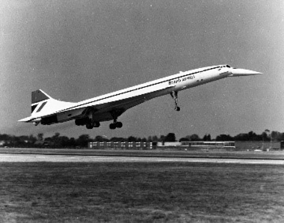 A black and white photo showing a large white jet plane with a distinctly turned down nose section in the process of taking off. Its landing gear is down and the words British airways are visible on the fuselage. Some low buildings are visible in the background. 