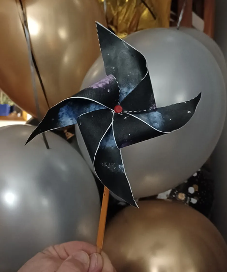 A hand is holding a pencil with a paper pinwheel with a galaxy design; balloons are visible in the background.