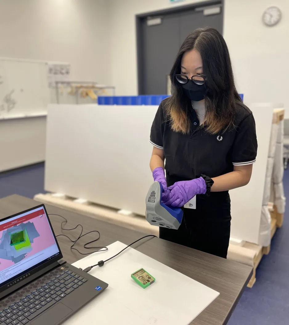 Woman with long, dark hair and wearing glasses, COVID-19 face mask, and gloves, stands at a table while holding a blue and grey 3D scanning device in front of a small, green dosimeter. A laptop on the table displays the dosimeter in 3D.