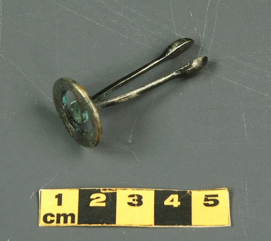 A small metal contraceptive, with a flat, open circular base and two parallel arms, joined at the base, with halved oval heads. This artifact is 5cm in length.