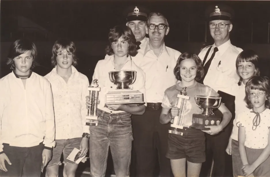 Une photographie noir et blanc montrant des filles et des garçons; deux des enfants tiennent des trophées. Trois hommes se tiennent en arrière-plan.