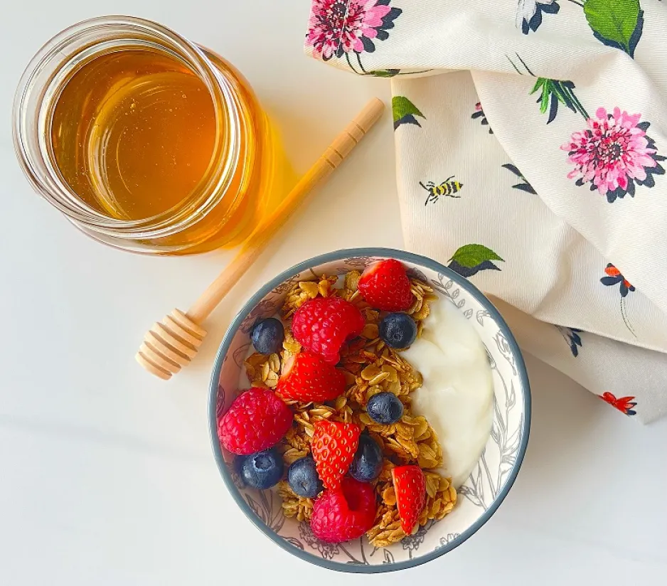 Un bol de granola avec des baies mélangées se trouve à côté d'un pot de miel ouvert, d'une cuillère à miel en bois et d'une serviette à fleurs.