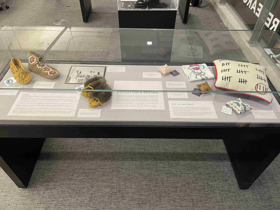 An exhibition display case. There are four moccasins on the left side of the case. On the right side is a pillow decorated with tally marks, beside which are other unidentified textile objects.