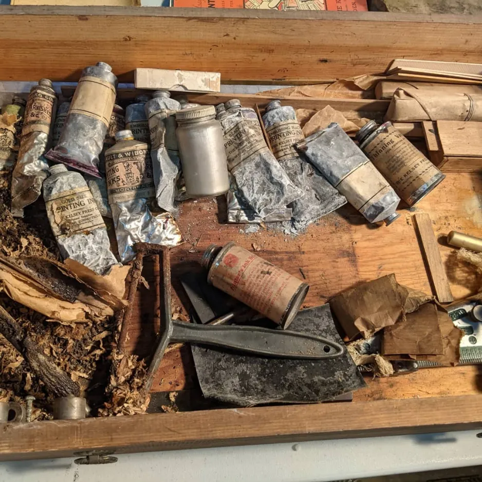 A wooden drawer filled with printmaking tools that are in various stages of degradation. Includes a mouse nest in the bottom left corner, several flaking lead ink tubes, and several packages of lead type wrapped in brown paper. 