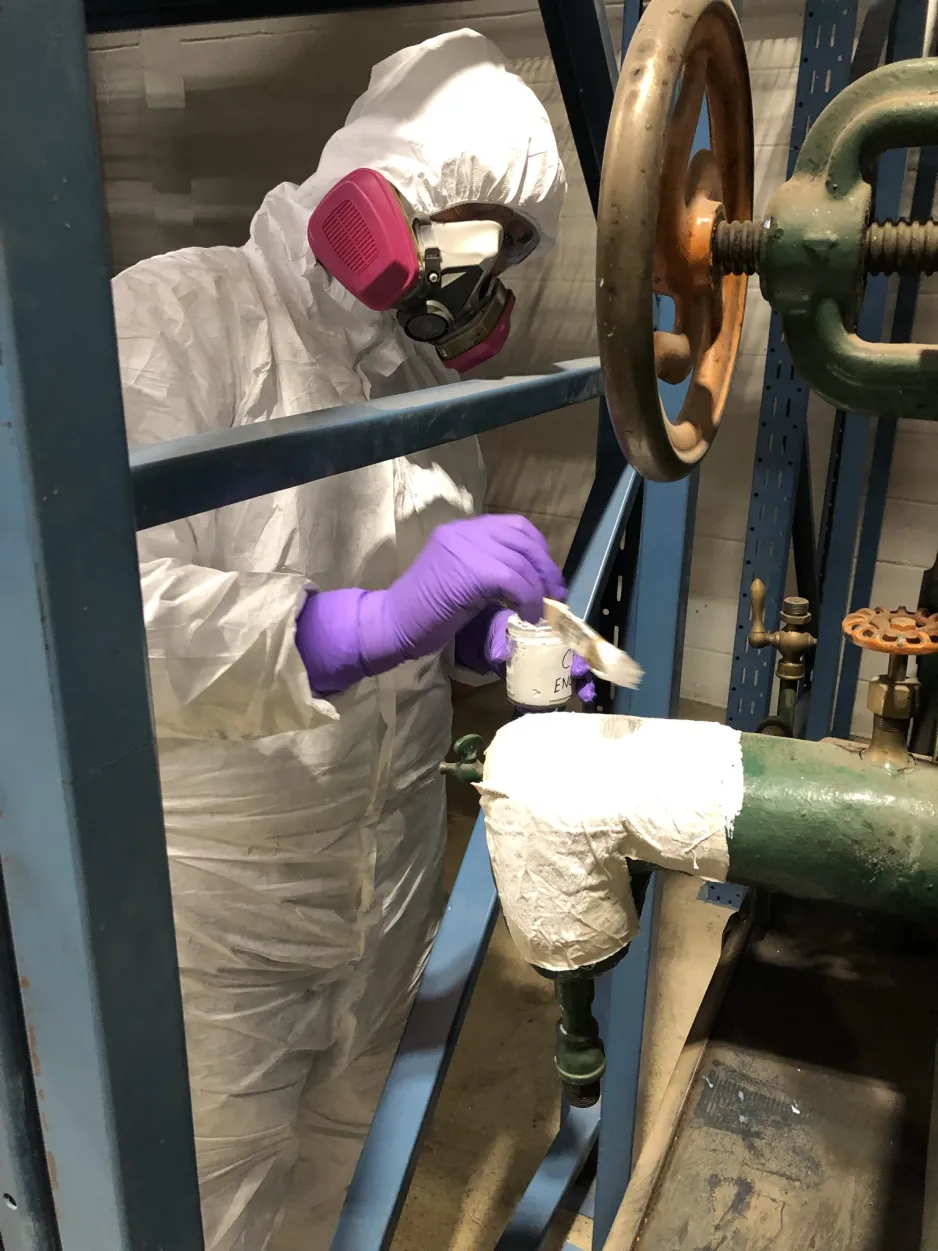  A person wearing a white protective suit, a ventilator, and purple gloves applies sealant to the asbestos insulation jacket of an artifact with a brush.