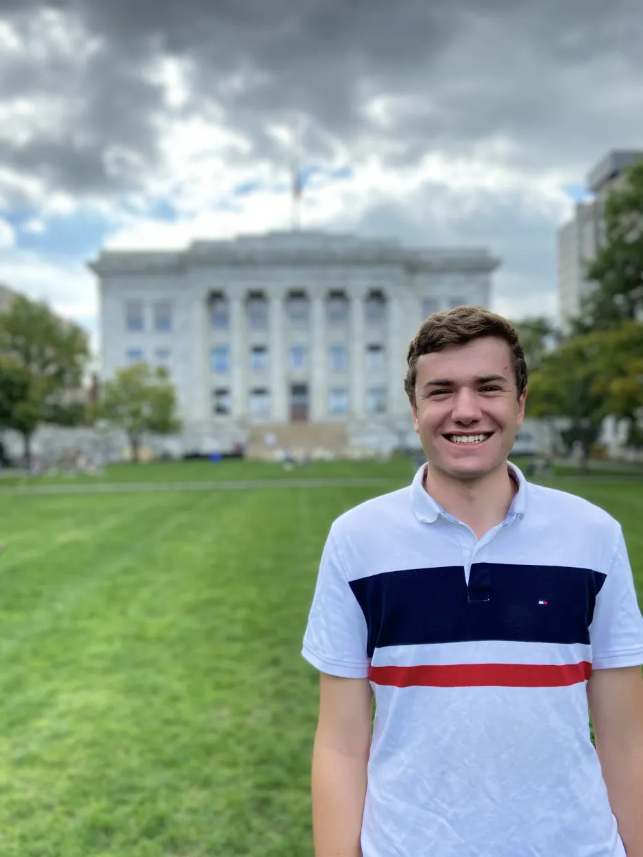 In the foreground, award winner Jackson Weir smiles directly at the camera. In the background, a grassy lawn and an impressive stone building are blurred but visible.