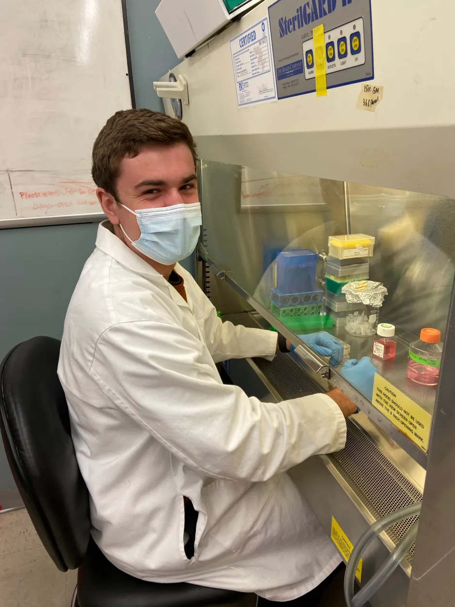 Award winner Jackson Weir wears a white lab coat, mask, and rubber gloves as he sits at a laboratory counter; his gloved hands are underneath a clear partition and there are several bottles and boxes visible.