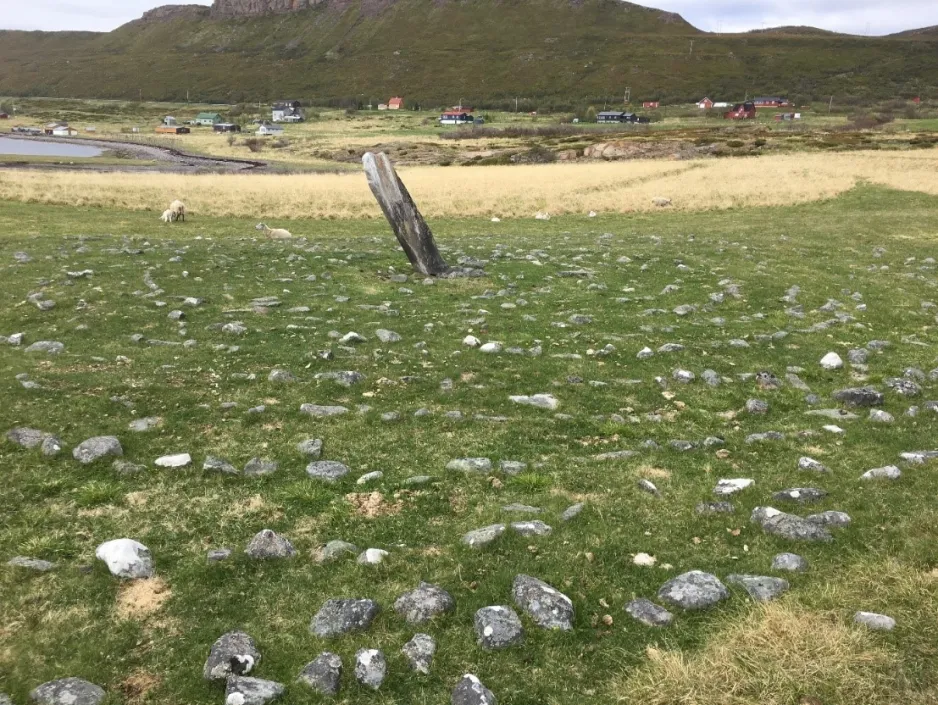 Image d’une structure traditionnelle en pierres dans un champ en Norvège, avec quelques petites maisons et une colline en arrière-plan.