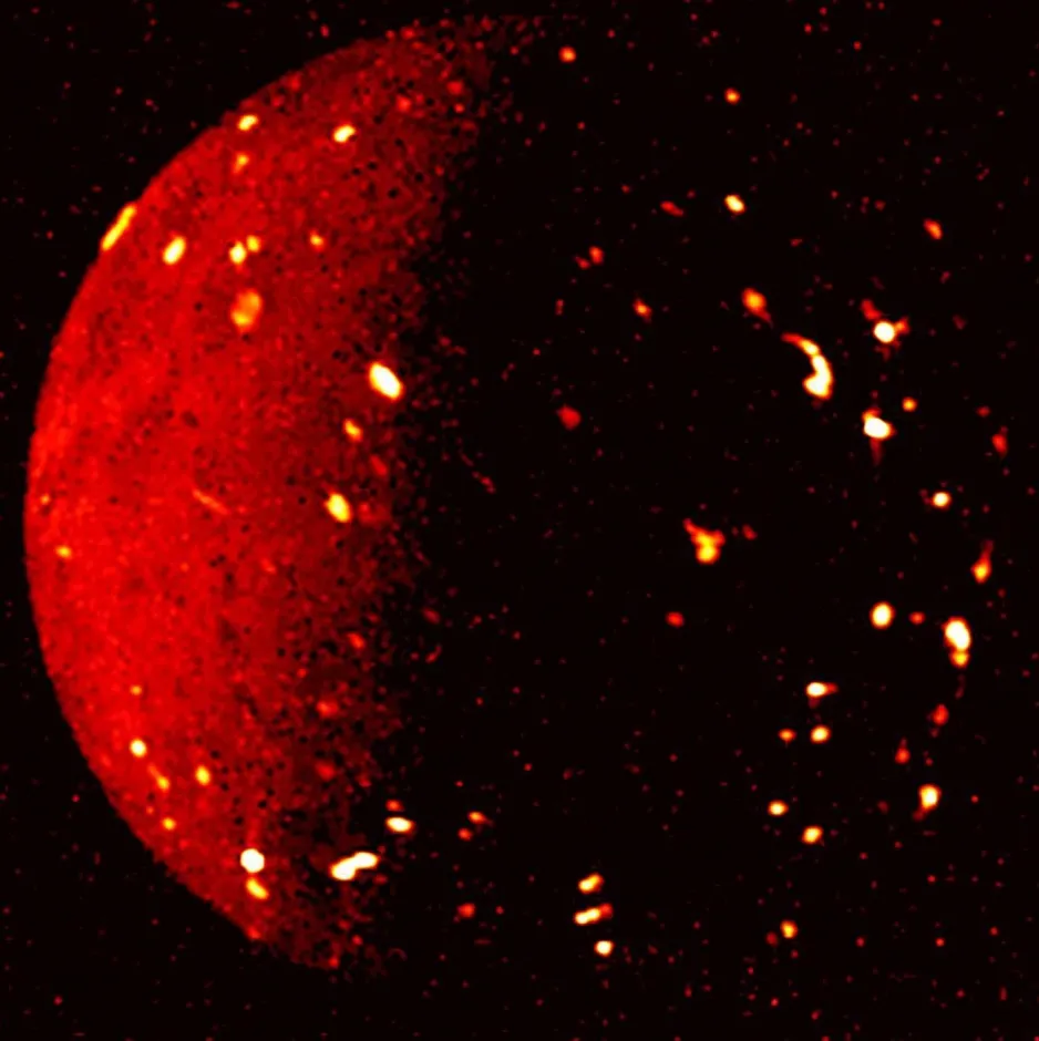 Une vue globale de Io en infrarouge. Le côté gauche est illuminé par le soleil et le côté droit est dans l’ombre, et le globe en entier est parsemé de taches claires représentant des volcans actifs.