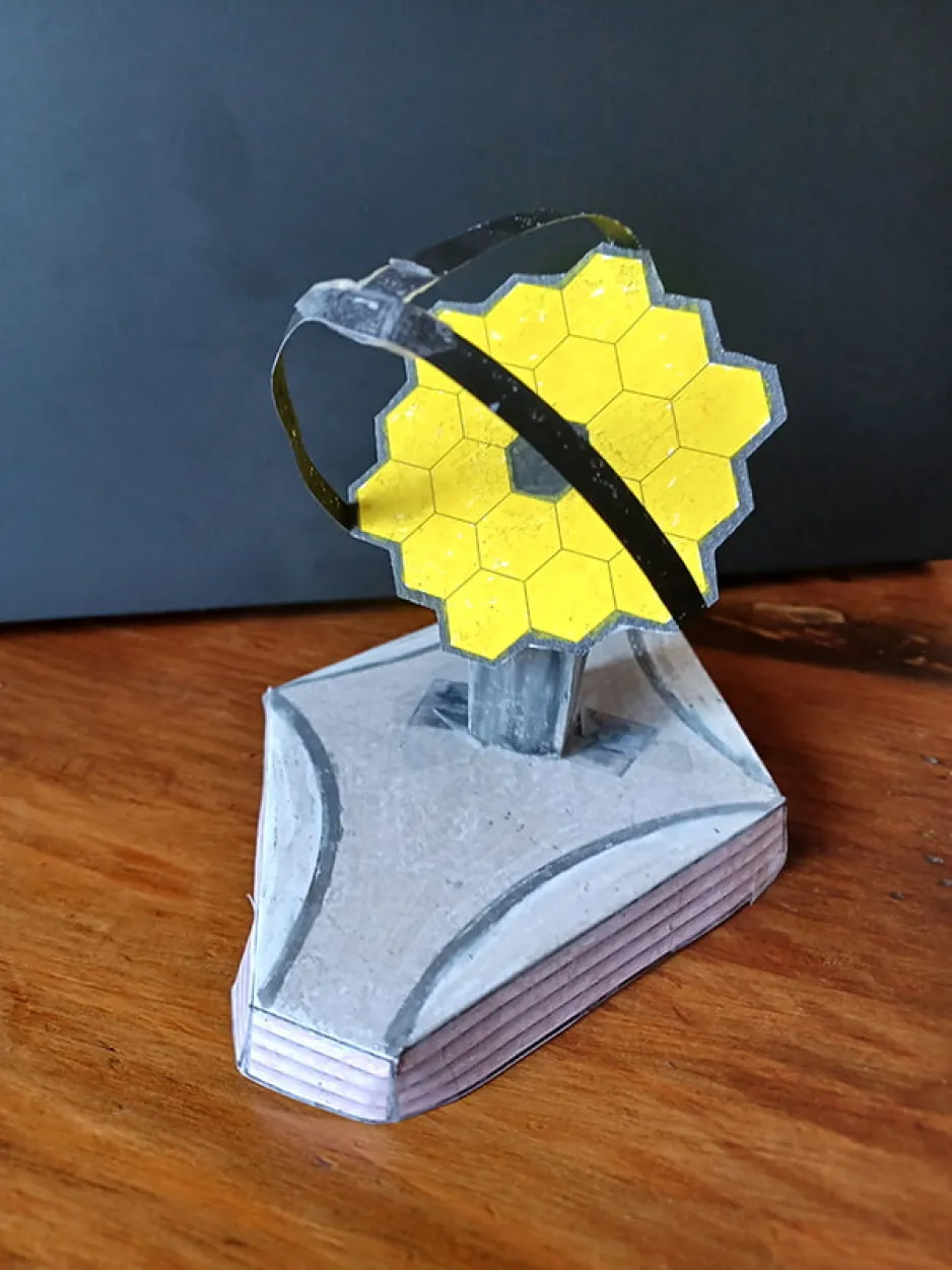 A small, paper model of the James Webb Space Telescope sits on a tabletop.