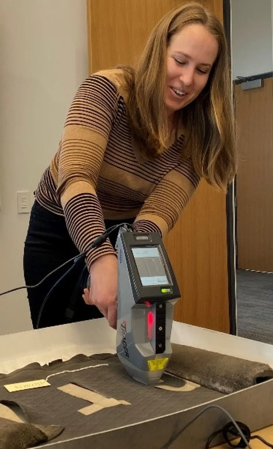A white woman wearing a striped sweater and holding an XRF that looks like a Star Trek phaser gun. The instrument head is resting on the surface of a rolled black artifact with large white lettering, and the letter “T” is visible.