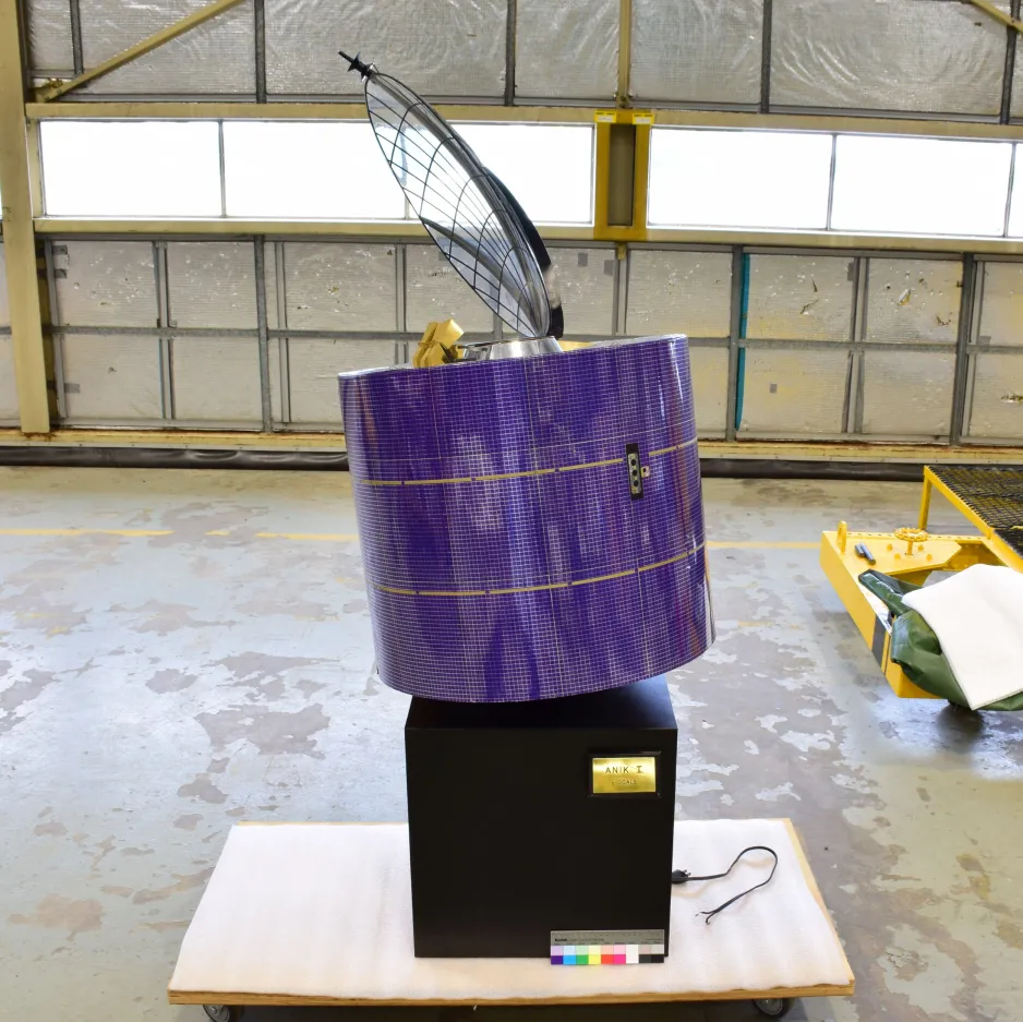 The satellite model sits on a wheeled pallet in a warehouse. It is purple with a slight checkered appearance and two yellow horizontal lines near the center. On top of the cylinder is a clear concave circle that curves towards the left side of the screen. The whole satellite is on top of a black box with a gold plaque that reads "ANIK I 1/3 SCALE". 