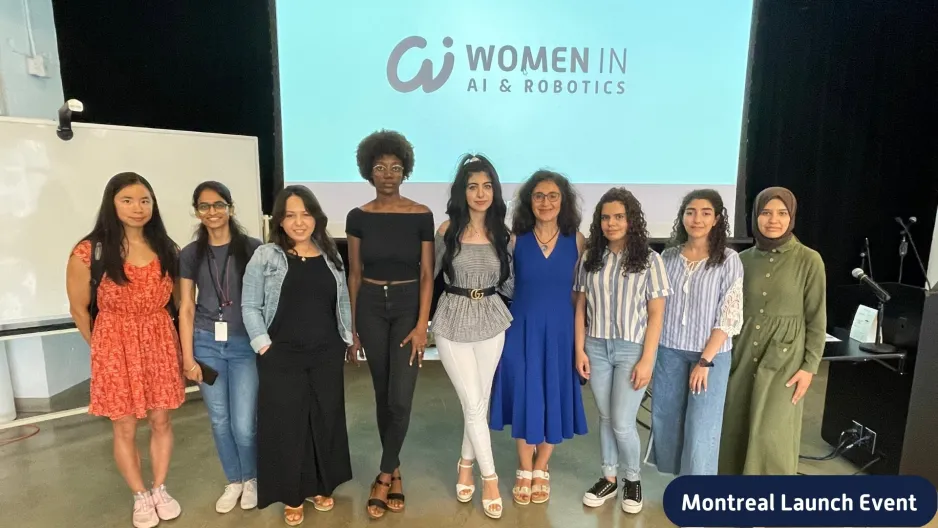 Nine women of various race stand in a line in front of a projector screen that reads "Women in AI and Robotics"