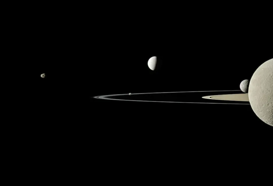 The rings of Saturn slice horizontally, almost edge-on, through the middle of the image. A variety of Saturnian moons of varying apparent sizes are in the image ranging from very small, background moons to larger and closer moons.