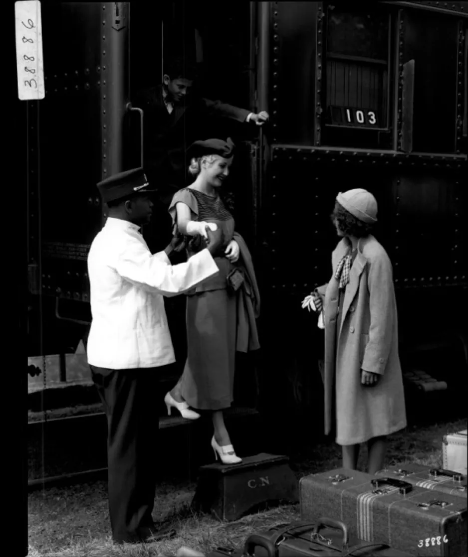 Black and white photograph of a Black porter helping a white woman disembark from the train. Behind the women stands a child at the top of the train steps. At the bottom right of the photograph another white woman waits for the women is disembark, luggage stands nearby.