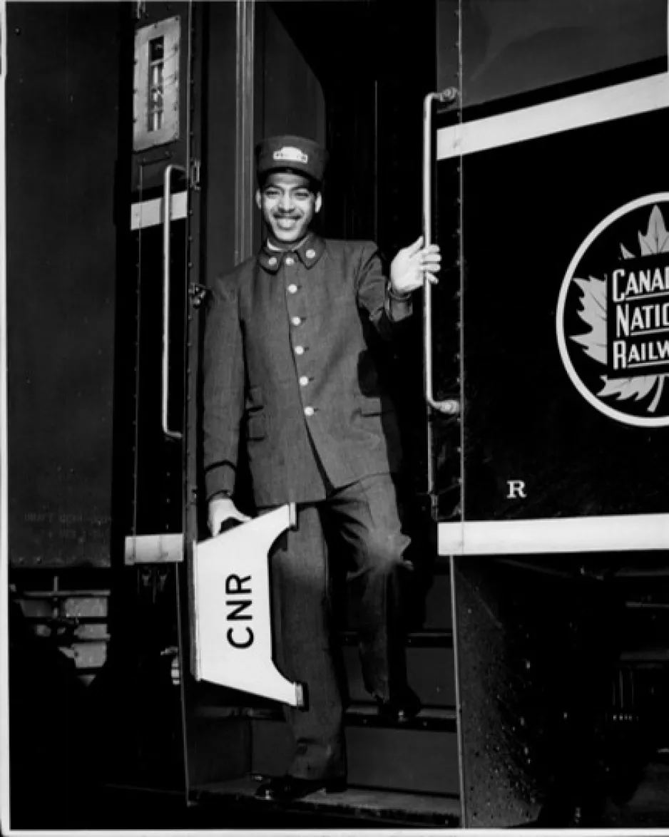 Black and white photograph of a Black porter smiling on train steps holding a step stool with the letters CNR on it.