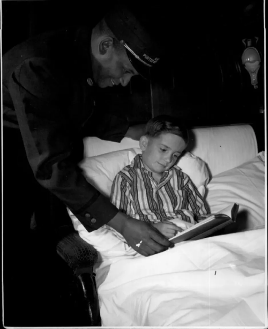 Photo noir et blanc d’un enfant blanc dans un lit, un livre à la main, et un porteur noir se penchant au-dessus de l’enfant pour l’aider.