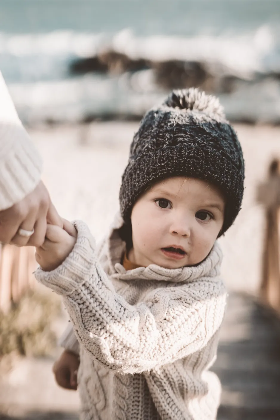 A toddler wearing a wool hat and wool sweater holds a grownup’s finger. There is a beach in the background. 