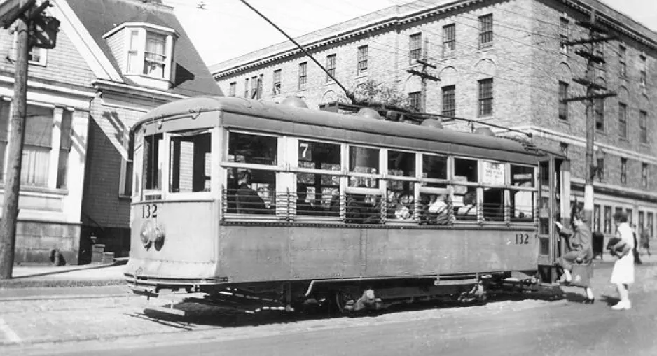 A black and white photograph of a trolley car stopped on a city street. Two passengers board the car at the rear.