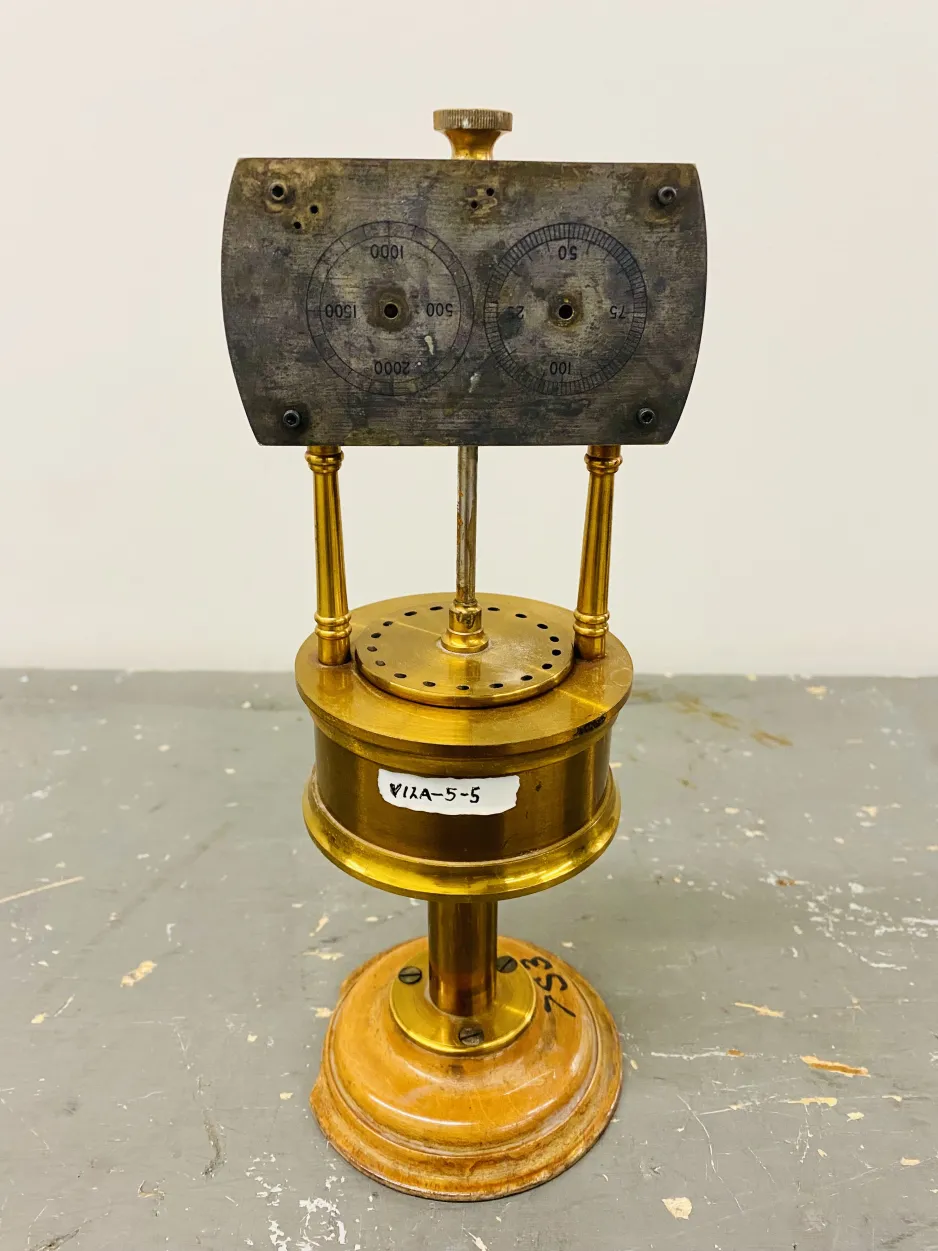 Full frame shot of Cagniard Siren. On top is an aged, rectangular metal panel, with circular gauges etched into the panel. On the bottom is a brass stand with a cylindrical unit in the centre, with holes punched into the top of its perimeter.
