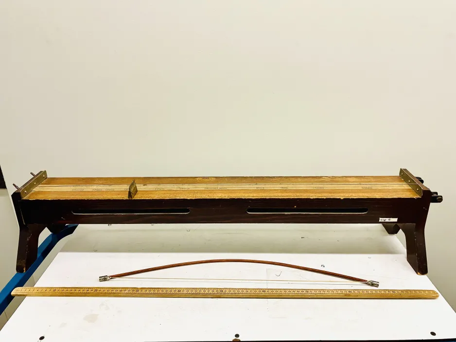 Full frame shot of a sonometer. The surface of the instrument is a rectangular piece of wood, positioned like a table, with a light-coloured centre strip running left to right across its length. There are two support structures, on the left and right, made of dark-coloured metal. A wooden bow is placed in front of the instrument.