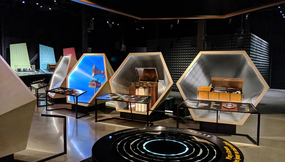 A wide shot of the Sound by Design exhibition taken from visitor’s perspective; several musical artifacts are visible inside hexagonal display cases.