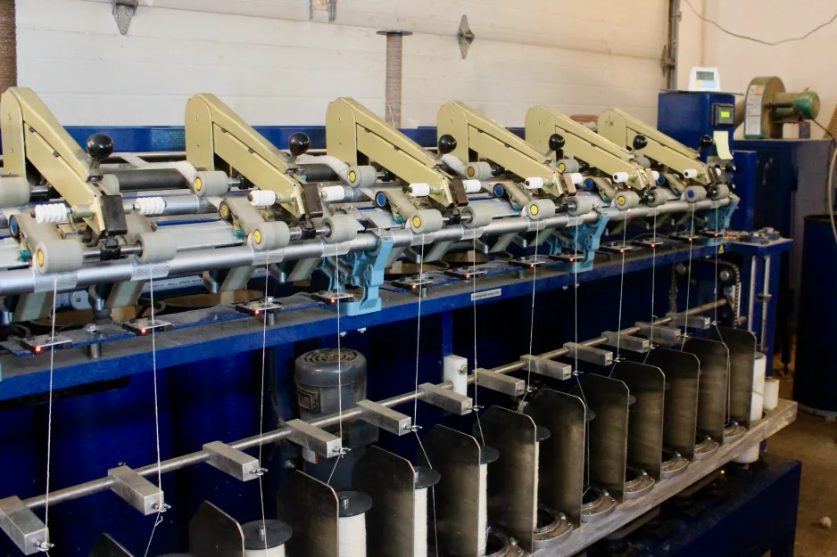 La fileuse de la ferme et filature Long Way Homestead est une machine industrielle de couleur bleue. Les cordons de laine, plus épais et pelucheux, sont tirés à travers une série de mécanismes de filage pour être tordus sous forme de fil. Cette machine file plusieurs fils de laine à la fois.