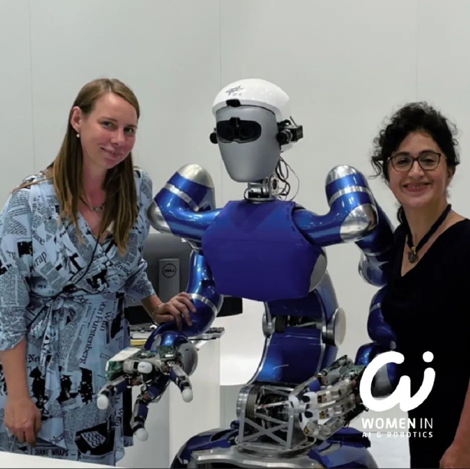 Two women stand on either side of a blue and silver, humanoid robot