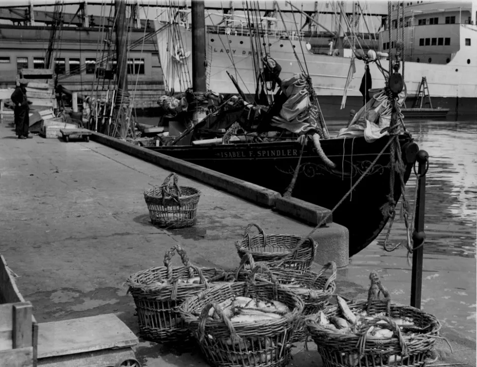 Black and white photograph with baskets of fish in the foreground. A ship with the word “Isabel F. Spindler” is docked in the mid-ground, with sails lowered. In the background a much larger ship with the words “Lady Drake” is docked beside a building.