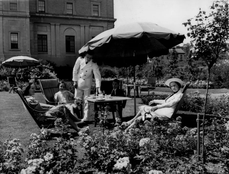 Une photo en noir et blanc de deux femmes et d’une fille assises dans des chaises de jardin inclinables sous un parasol. Un serveur vêtu d’un uniforme blanc met la table pour le thé. La scène est encadrée de rosiers.
