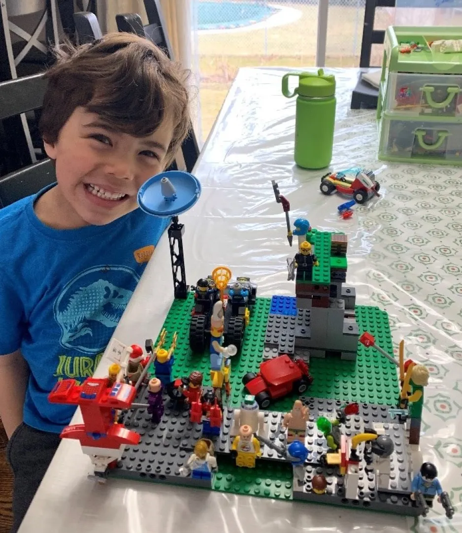 Un petit garçon vêtu d’un t-shirt bleu assis près d’une table affiche un grand sourire. Sur la table, on voit une création colorée complexe en LEGO.