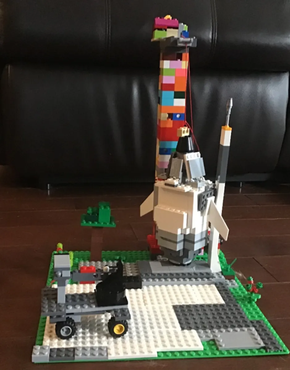 Une création LÉGO qui inclue une fusée, une tour et une rampe de lancement, et un véhicule lunaire. La création est debout sur un plancher de bois avec un arrière-plan noir.