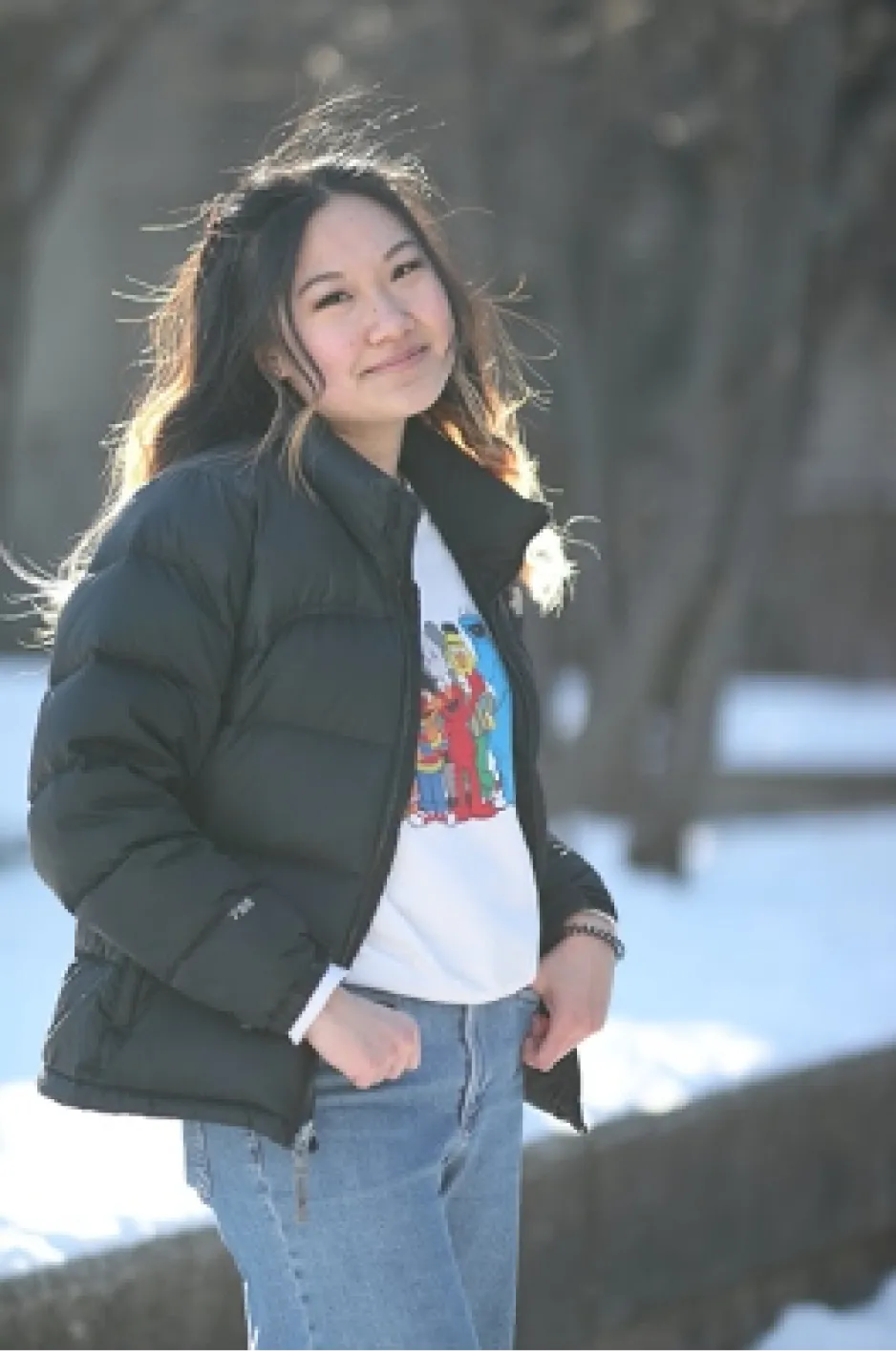 A young woman with pale skin, long, black hair with orange-blonde ends, wearing a black puffer jacket, a white sweatshirt with Sesame Street characters, and blue jeans smiling against a snowy winter background.
