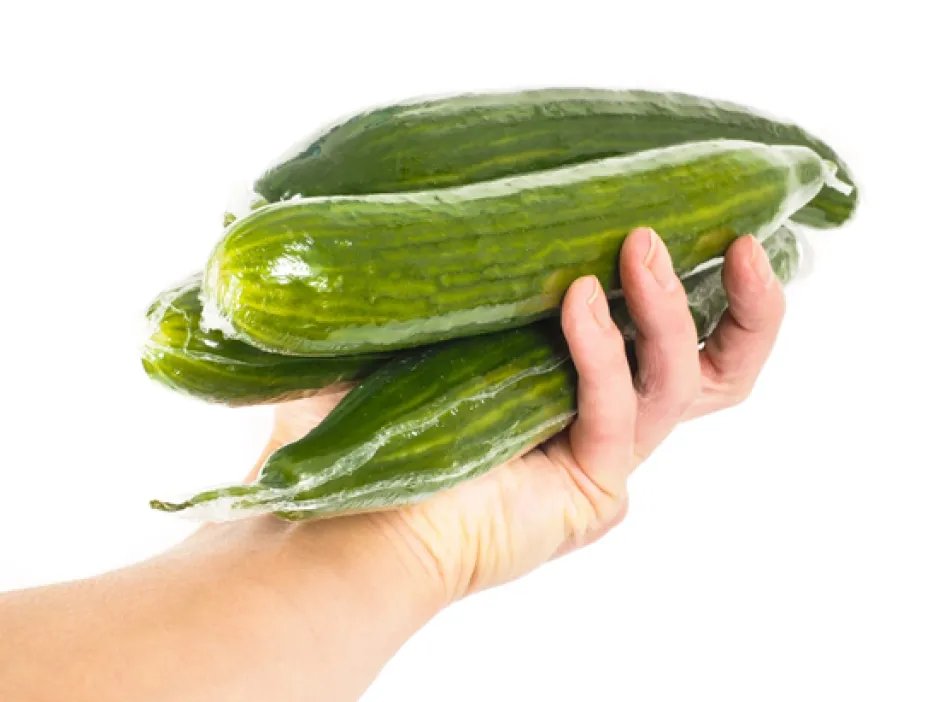 An outstretched hand holding four English cucumbers in clear plastic shrink wrap.
