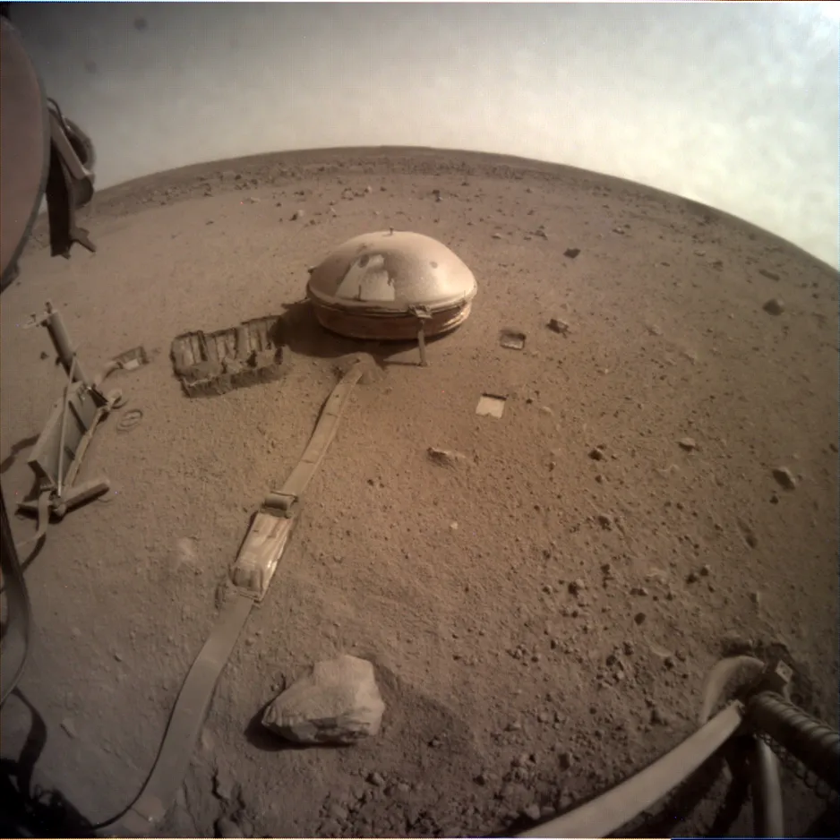 A desert terrain centred on a tethered, dusty white dome (seismometer) with small scoop marks in the sand on the left. The lander’s foot is on the right.