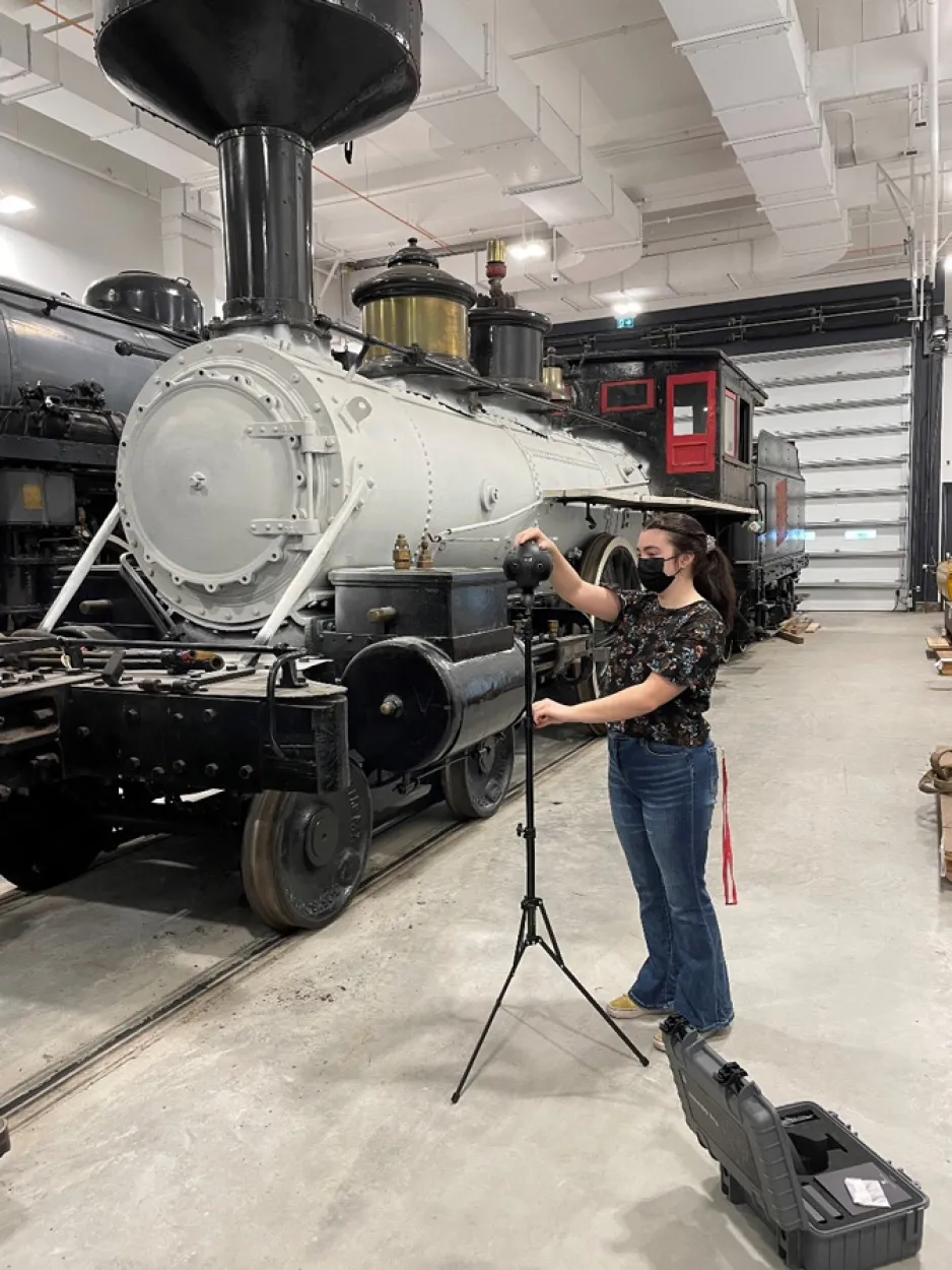 A woman standing beside a spherical camera on  a tripod, with a large locomotive in the background.