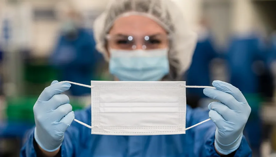 A woman wearing personal protective equipment holds a white face mask at arm’s length; she is blurred in the background and the mask is in focus.