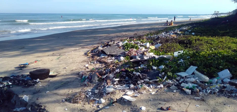 An ocean beach with piles of trash along the shore. Four people are sitting on the beach in the distance.