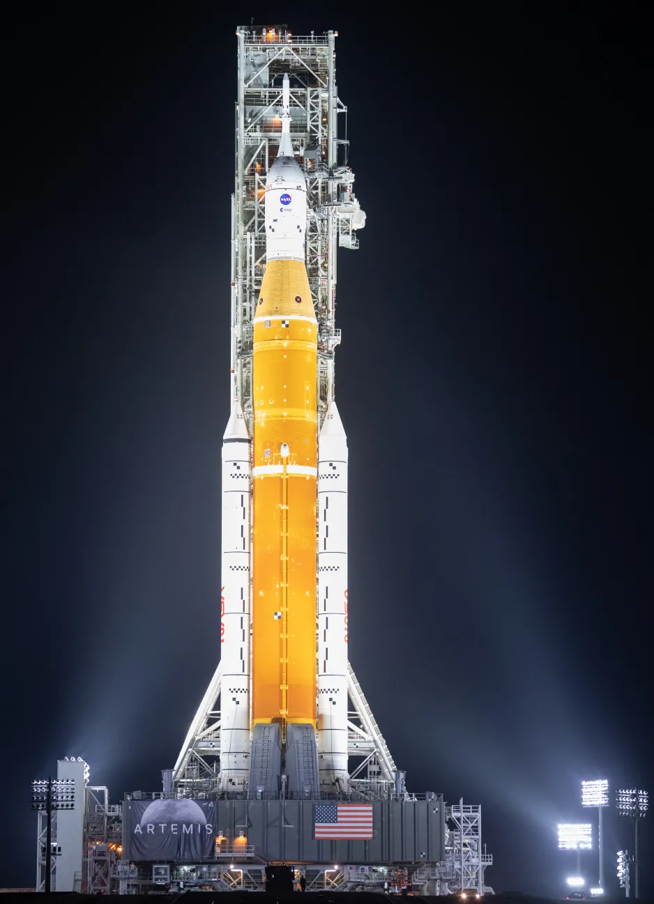 Un énorme noyau de fusée orange en position verticale entre deux propulseurs de fusée sur un véhicule de lancement mobile, éclairé par des projecteurs sur un fond noir