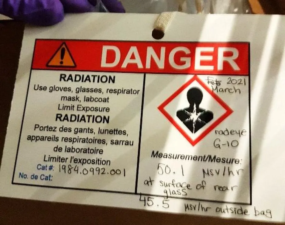 A white paper hazard tag with black writing, a red “DANGER” banner, and the WHMIS health hazard pictogram. The tag has the following printed text: “RADIATION Use gloves, glasses, respirator mask, labcoat Limit Exposure”, and the handwritten text: “2021 March, radeye G-10, 50.1 µSv/hr at surface of rear glass, 45.5 µSv/hr outside bag, 1984.0992.001”.