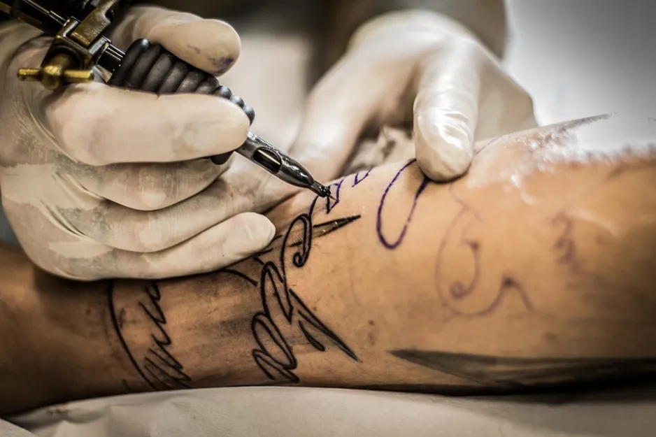 A close up of a forearm being tattooed. The person being tattooed has some drawings on their skin being traced by the tattoo artist. The forearm already has black tattoos. The tattoo artist is wearing white medical gloves and holding a tattoo gun. 
