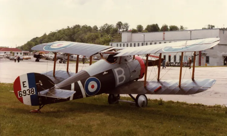 A small biplane, photographed outdoors from the back right, with a light gray utilitarian building in the background.