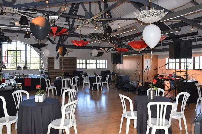 A large room with industrial-chic barn-style dark metal beams on a high and bright ceiling. The floor and walls are wood-toned and the back of the room has multi-coloured floor to ceiling glass panes and semi-circle windows. Small blue tables are set up around the room and four white chairs are at each one. Helium balloons float above each of the tables.