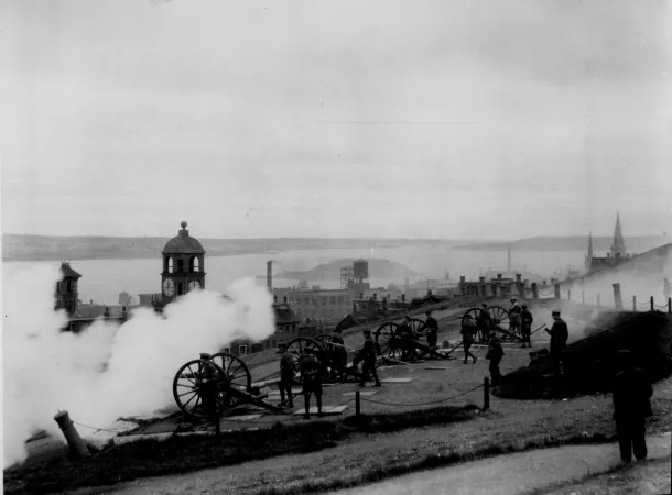 Une photo en noir et blanc de soldats debout à côté d’une série de canons sur une colline. Il y a de la fumée dans les airs, indiquant que les canons viennent tout juste d’être tirés. Le havre d’Halifax est visible en arrière-plan.