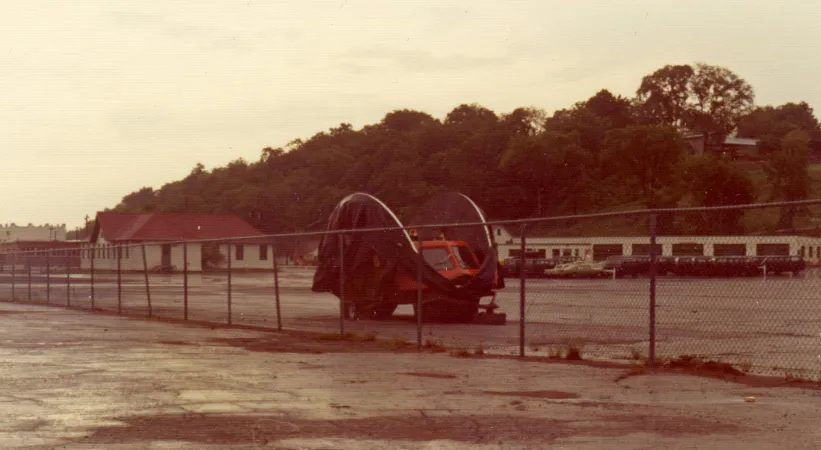 An odd-looking vehicle is sitting outdoors, behind a chain-link fence. The vehicle has black semi-circular sides that protrude above its orange body. A two-pane windshield seems to peek out of its folded structure.